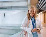 Uśmiechnięta studentka medycyny w białym kitlu, z plecakiem i stetoskopem, trzyma tablet. Zastanawia się, gdzie najłatwiej dostać się na medycynę.