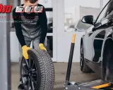 Mecánico con guantes amarillos sosteniendo una rueda, listo para cambiar neumáticos. El coche está en un taller.
