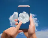 Dłonie trzymają telefon, na którego ekranie widać chmurę. To symbol tego, gdzie jest chmura w telefonie - w chmurze danych.