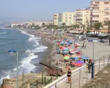 Sonniger Torrox Strand mit vielen bunten Sonnenschirmen, Menschen im Meer und an Land, sowie historischen Kanonen.