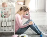Zmartwiona mama siedzi obok łóżeczka, zastanawiając się, depresja poporodowa ile trwa. Dziecko bawi się w łóżeczku.