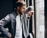 Mężczyzna zamyślony przy oknie, trzymający szklankę z bursztynowym płynem. Czy to jeden z ukryty alkoholizm objawy?