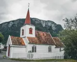 Biały kościółek z czerwoną wieżą i dachem, otoczony białym płotkiem, na tle surowych gór. Symbol norweskiej religii i spokoju.