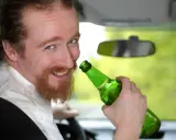 Uśmiechnięty mężczyzna z rudą brodą trzyma zieloną butelkę piwa w samochodzie. Mówi, że piwo to nie alkohol, ale jazda z nim jest niebezpieczna.