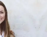 Lächelnde Frau mit sehr dicken Haaren und einem beigen Mantel vor einer hellen Wand.