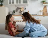 Madre e hija ríen en el sofá. Ella le enseña como es la primera regla de una niña: la empatía.