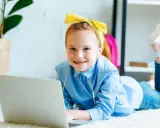 Uśmiechnięta dziewczynka w żółtej opasce i niebieskiej koszuli leży na dywanie z laptopem. Słucha muzyki przez słuchawki, jak wiele dzieci.