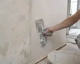 Ręka trzymająca kielnię wygładza tynk na ścianie. Widać starą, łuszczącą się farbę obok.