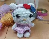 Muñeca de Hello Kitty tejida a crochet, con vestido lila y lazo rojo y azul. Hilos y agujas de crochet alrededor, perfectos para crear tus propios hello kitty crochet patrones español.