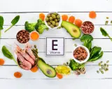 Wokół tabliczki z napisem "Witamina E" ułożono produkty bogate w ten składnik: krewetki, migdały, orzeszki ziemne, awokado, oliwki, brokuły, suszone morele, szpinak i olej.
