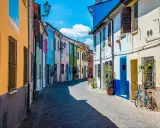 Un vicolo colorato a San Giuliano, con case dipinte di giallo, blu, verde e rosa. Una bicicletta azzurra è parcheggiata davanti a una casa.