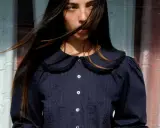 A young woman with windswept hair wears a dark blue blouse with a charming peter pan neckline, delicate embroidery, and puffed sleeves.