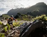 Läufer kämpft sich auf Madeira bergauf, vorbei an Felsen und gelben Ginsterbüschen, mit Wolkenmeeren und schroffen Gipfeln im Hintergrund.