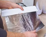 Ręce trzymają biały dozownik z folią aluminiową. Zastanawiasz się, która folia aluminiowa strona do pieczenia? Ta błyszcząca strona odbija ciepło.