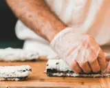 Hände mit Handschuhen rollen Sushi auf einem Holzbrett. So einfach ist Sushi selbst machen!