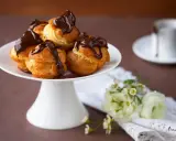 Un plateau blanc présente des profiteroles gastronomiques garnies de crème et nappées de chocolat noir. Des fleurs délicates ajoutent une touche de fraîcheur.