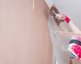 Ręce w czerwonych rękawicach nakładają obrzutkę cementową na ścianę za pomocą kielni.
