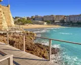 Paseo marítimo de madera junto a rocas y mar turquesa, con playa y edificios al fondo. Un tramo del camí de ronda Tarragona.