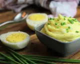 Cremige Sauce Tartar, garniert mit Schnittlauch, neben halbierten Eiern auf einem Holzbrett.