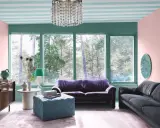 Ett vardagsrum med art deco inredning, mjuklila soffa, mörkgrå soffa, turkos pall och en stor fönsterutsikt över skogen.