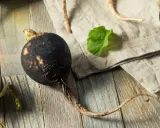 Gros radis noir rustique, avec ses racines fines, posé sur une planche de bois vieillie. Une feuille verte et un tissu de lin complètent la scène.