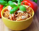 Pyszne spaghetti bez mięsa w żółtej misce, udekorowane bazylią i parmezanem. Obok pomidory i papryka.