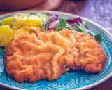 Ein knuspriges, originales Wiener Schnitzel auf einem blauen Teller, serviert mit Kartoffelsalat und frischem Grün.