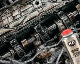 Widok wnętrza silnika z widocznymi wałkami rozrządu i śladami oleju. Obok butelka płukanki silnika K2 Motor Flush, sugerująca problemy po jej użyciu.