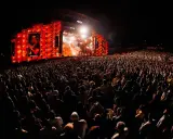 Un enorme pubblico al festival in Portogallo, illuminato da un palco con luci arancioni e un'immagine su schermo.