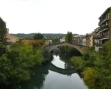 El puente de la cárcel de Estella cruza un río sereno, reflejando su arco de piedra entre frondosa vegetación y edificios históricos.