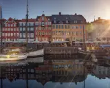 Färgglada hus vid en kanal i Köpenhamn, med båtar i förgrunden. Solen skiner och reflekteras i vattnet. En perfekt vy för en flyg till Köpenhamn-resa.