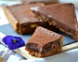 Tranche de tarte façon snickers, garnie de chocolat et de cacahuètes, saupoudrée de cacao. Une fleur bleue décore la présentation.