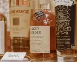 Flera flaskor whisky, inklusive Monkey Shoulder och Glenmorangie, står uppradade. En liten skylt med texten "Scotch" syns framför.