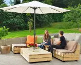 Två personer njuter av solen under ett parasoll vid en hemmabyggd loungesoffa i trä.