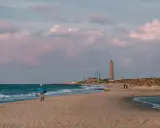 En person promenerar på stranden vid Skagens fyr. Molnen färgas rosa i solnedgången.