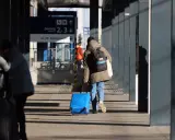Osoba z niebieską walizką na kółkach i plecakiem idzie po peronie.
