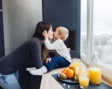 Matka całuje dziecko, siedząc przy oknie. Na blacie owoce i soki.