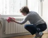 Mujer limpiando un radiador blanco con un paño rosa. Un ejemplo de cómo limpiar radiadores para mantener el hogar cálido y limpio.