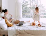 Kobieta gra na ukulele, a dziecko bawi się klockami na łóżku.