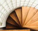 Escalera helicoidal de madera con peldaños que giran bajo un techo de bloques de vidrio translúcido.