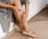 Mani che accarezzano le gambe, evidenziando la pelle secca. La persona è seduta su un letto, avvolta in un accappatoio grigio.