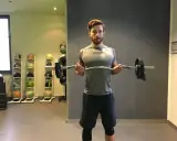 Mann macht Bizeps-Curls mit einer Langhantel. Im Hintergrund stehen Medizinbälle.