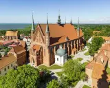 Zamek Frombork z katedrą i wieżami, otoczony zielenią, nad błękitną wodą.