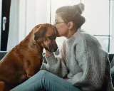 En kvinna kysser sin hunds huvud, en gest av saknad och sorg. Hunden ser trött ut, som om den delar hennes känslor.