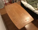 Ett slitet soffbord i trä, redo att måla om. Bredvid syns en säng med vitt och grönt överkast.