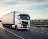 Biała ciężarówka jedzie autostradą w Holandii. W tle widać magazyny i kontenery.
