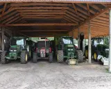 Kilka zielonych i jeden czerwony traktor stoją pod drewnianą wiatą. To pewnie ich tymczasowe schronienie, bo taka wiata bez pozwolenia to nie najlepszy pomysł.