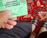 Zielona legitymacja ZUS "LEGITYMACJA EMERYTA-RENCISTY" w dłoni. Na dywanie widać stopy w kapciach.