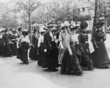 Frauen in langen Kleidern und Hüten marschieren entschlossen. Ein historisches Bild der feministischen Bewegung.