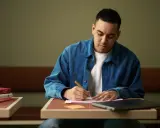 En ung man skriver i ett anteckningsblock, omgiven av böcker och en laptop. Bra ny på jobbet tips kan vara att anteckna flitigt.